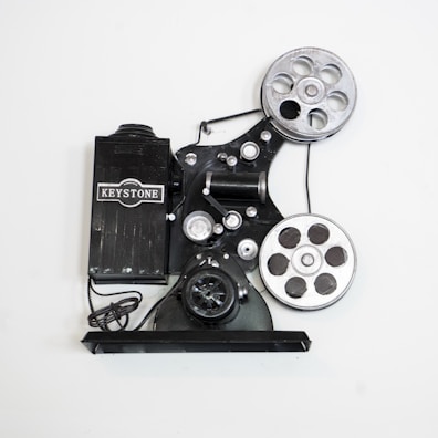 A vintage film projector with two large reels and various mechanical components. The device is marked with the brand name 'Keystone' and features a classic, industrial design with metallic and black parts.