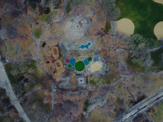 An aerial view of a park showcasing a playground area with various colorful, circular features, surrounded by trees and grassy areas. Pathways meander throughout the park, and there's a mix of barren ground and patches of vegetation. To the side, roads and a few vehicles are visible, indicating proximity to urban streets.