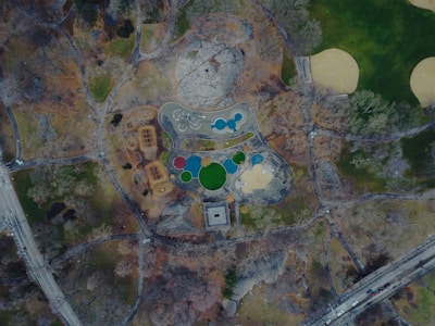 An aerial view of a park showcasing a playground area with various colorful, circular features, surrounded by trees and grassy areas. Pathways meander throughout the park, and there's a mix of barren ground and patches of vegetation. To the side, roads and a few vehicles are visible, indicating proximity to urban streets.