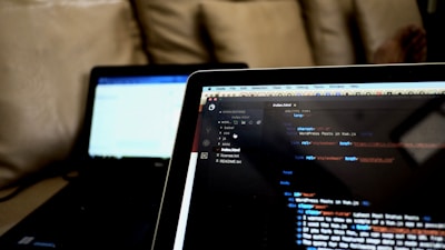 A close-up view of two laptops indoors, displaying a code editor with HTML code on the screen of the laptop in the foreground. The second laptop in the background is out of focus. Both laptops are placed on a cozy couch, creating a casual and comfortable working environment.