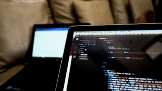 A close-up view of two laptops indoors, displaying a code editor with HTML code on the screen of the laptop in the foreground. The second laptop in the background is out of focus. Both laptops are placed on a cozy couch, creating a casual and comfortable working environment.