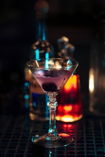 A cocktail glass filled with a purple beverage, garnished with a cherry, set on a bar surface. In the background, there are blurred bottles with colorful lights creating a warm and inviting atmosphere.