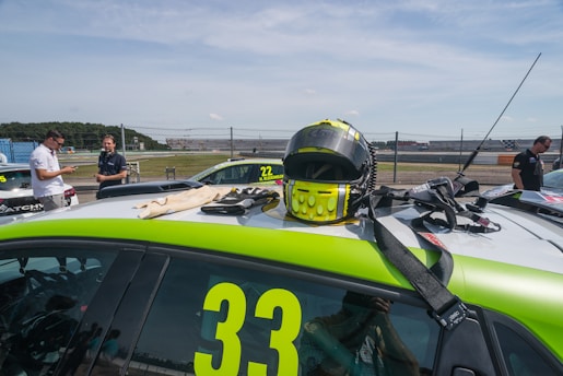 A racing helmet is placed on top of a green and white race car marked with the number 33. Nearby other cars in the background, marked with different numbers, are visible. Several people are standing around, some looking at their phones. The scene takes place in an outdoor racing track with a fenced boundary and a clear sky.