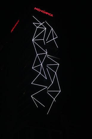 An illuminated building facade featuring white geometric LED light patterns against a dark background. The design appears to be abstract with intersecting lines and angles. The brand name Movenpick is visible at the top in bright red letters.