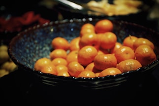 A bowl filled with numerous small, bright orange fruits, likely tangerines or mandarins, set against a dark background. The bowl has a textured, blue interior that contrasts with the vivid color of the fruit.