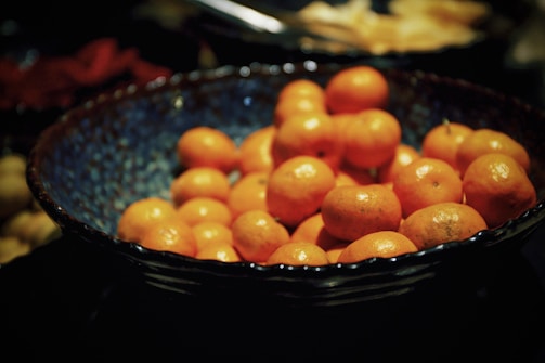 A bowl filled with numerous small, bright orange fruits, likely tangerines or mandarins, set against a dark background. The bowl has a textured, blue interior that contrasts with the vivid color of the fruit.