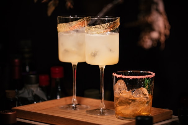 Two tall glasses filled with a pale yellow cocktail have rims decorated with a golden sugar coating, while a shorter glass with a golden drink features pink sugar on the rim and contains large ice cubes and rosemary garnish. The scene is set on a wooden tray against a dark background with dim lighting.