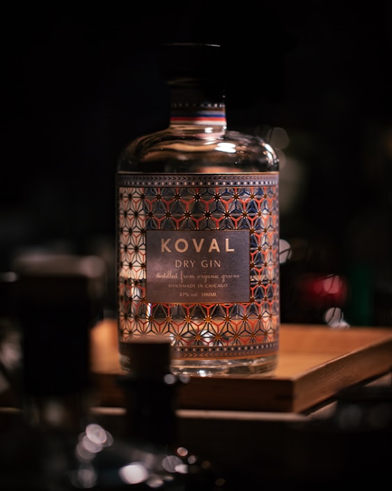 A glass bottle of Koval dry gin is prominently displayed, featuring an ornate geometric design on its surface. The label reads 'Koval Dry Gin,' noting that it is 'distilled from organic grains' and 'handmade in Chicago.' The bottle sits on a wooden platform with a softly blurred background, creating a warm, luxurious atmosphere.