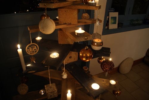 A handcrafted wooden tree structure is adorned with various decorative ornaments and candles. The ornaments include metallic spheres, intricate wooden carvings, and geometric shapes. Candles are strategically placed on different levels of the tree, illuminating the scene with a warm, soft glow.