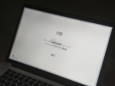 A laptop screen displaying a motivational quote in black text on a white background. The quote reads 'You will succeed because most people are lazy.' The screen is partially visible with the laptop keyboard and body in view.