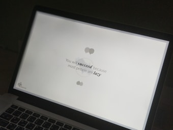A laptop screen displaying a motivational quote in black text on a white background. The quote reads 'You will succeed because most people are lazy.' The screen is partially visible with the laptop keyboard and body in view.