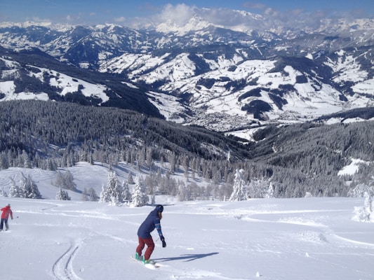 A winter landscape featuring snow-covered mountains and pine trees. In the foreground, a person is snowboarding down a pristine, untouched slope, with another person walking in the snow. The background reveals a vast, snowy valley with mountainous terrain under a partly cloudy sky.