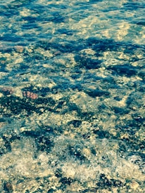 The image captures the texture and movement of transparent water flowing over a rocky surface, with various sizes and shapes of stones visible underneath the water. Sunlight reflects off the water's surface, creating shimmering patterns.