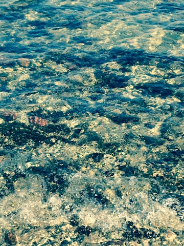 The image captures the texture and movement of transparent water flowing over a rocky surface, with various sizes and shapes of stones visible underneath the water. Sunlight reflects off the water's surface, creating shimmering patterns.