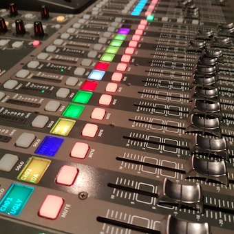 A close-up view of an audio mixing console featuring multiple rows of sliders, knobs, and brightly lit buttons in various colors such as red, green, blue, and yellow. Each slider is marked with numerical values, and labels indicate channels or settings. The surface is metallic with a professional, high-tech appearance.