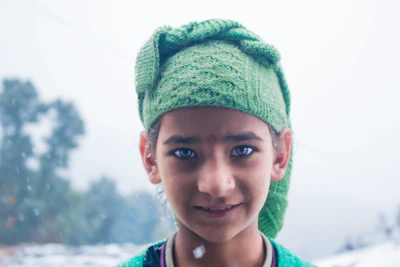 A child wearing a sage green crocheted hat, smiling outdoors with soft terracotta leaves around.