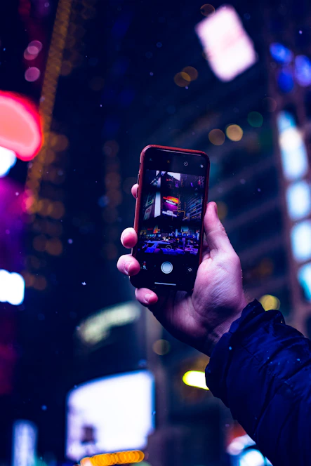 A cheerful young woman using a smartphone outdoors, capturing a photo with a bright city background.