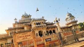 An intricately designed historical building with domes and decorative arches, set against a clear blue sky. Birds are flying around the structure, and a sign in the foreground reads 'Central Museum Jaipur'. The building has detailed architectural elements typical of Indian heritage.