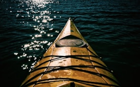A kayak is seen from the viewpoint of the person inside, with its tip cutting through the dark, rippling water. Sunlight reflects off the water, creating a sparkling effect.