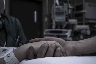 A hand lightly grasps another hand in a hospital setting. The scene includes medical equipment in the background and both hands suggest a supportive gesture. The person in the hospital bed is wearing a patient wristband.