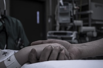 A hand lightly grasps another hand in a hospital setting. The scene includes medical equipment in the background and both hands suggest a supportive gesture. The person in the hospital bed is wearing a patient wristband.