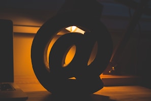 A large, three-dimensional at (@) symbol is silhouetted against a warm, glowing light source. The foreground is a deep shadow, casting elegant shapes on a wooden surface. The lighting creates a cozy and inviting ambiance, with glimpses of a desk lamp and a laptop in the background.