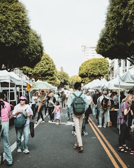 A busy street market with people walking and interacting among stalls covered with white canopies. Tall trees line both sides of the street, creating a picturesque scene. Adults and children are visible, some carrying bags and others pushing strollers.