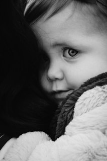 A close-up black and white portrait of a young child, partially hidden behind someone, with wide eyes and a curious expression. The child is wrapped in a soft blanket, giving a cozy and intimate feel to the image.