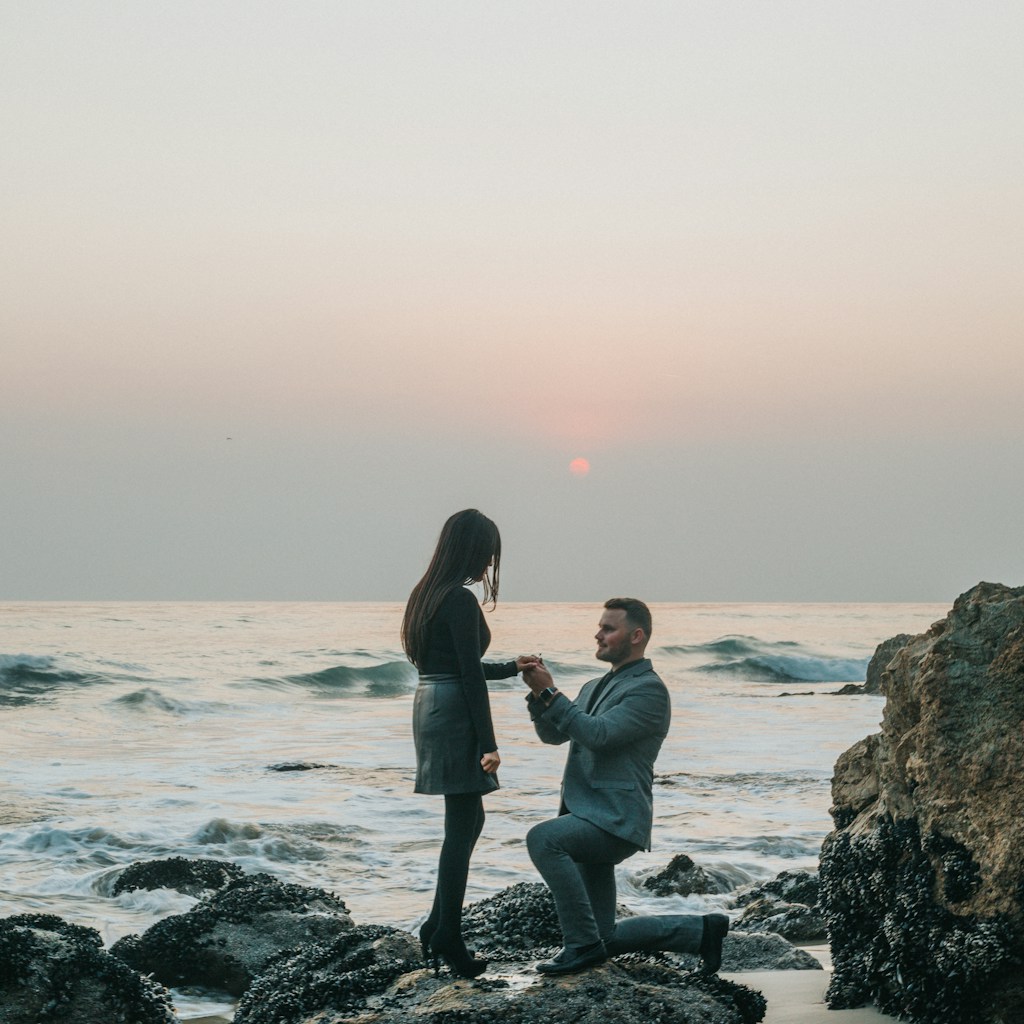 man kneeling in front of woman on rock at beach