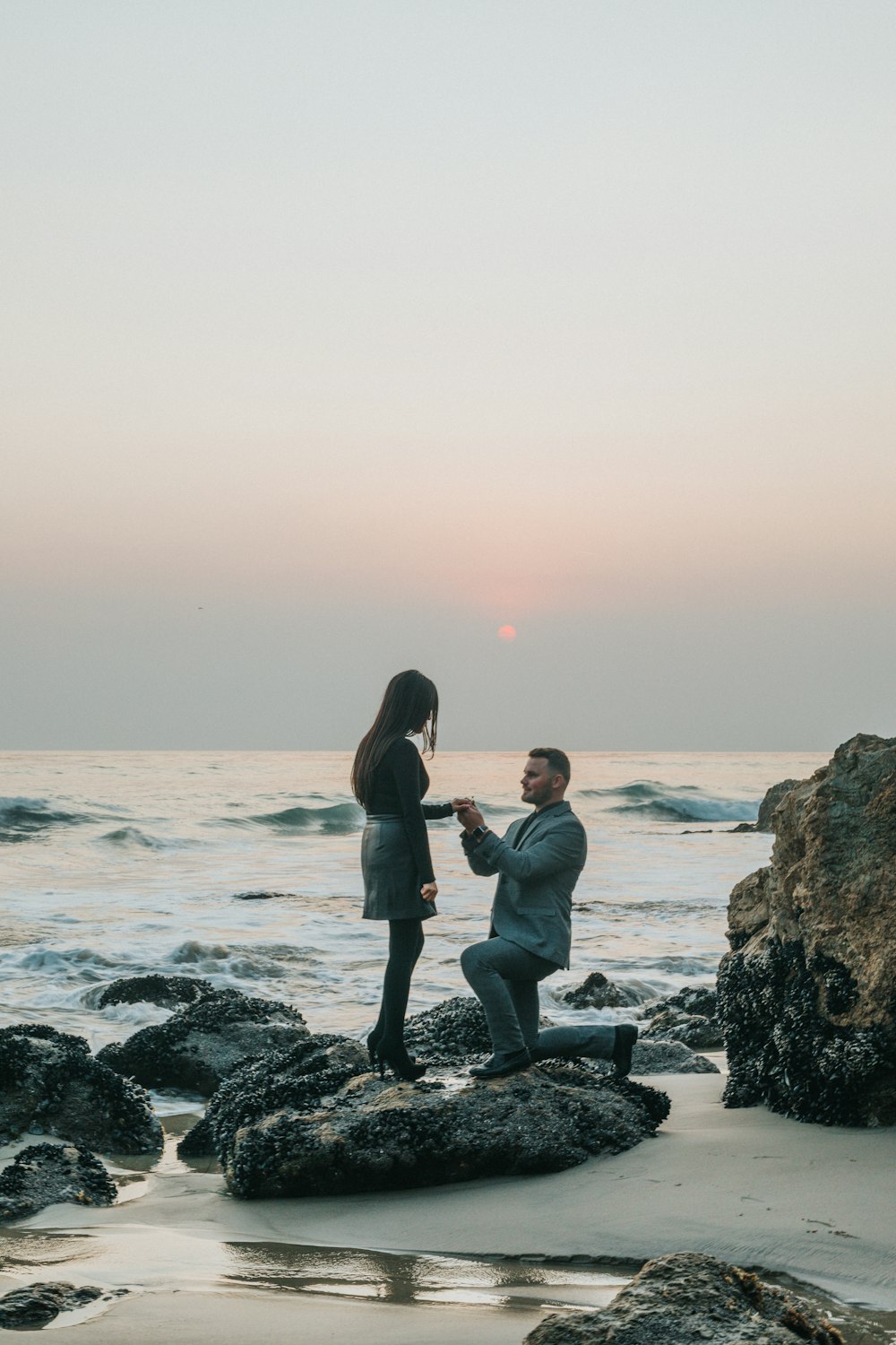 Mann kniet vor Frau auf Felsen am Strand