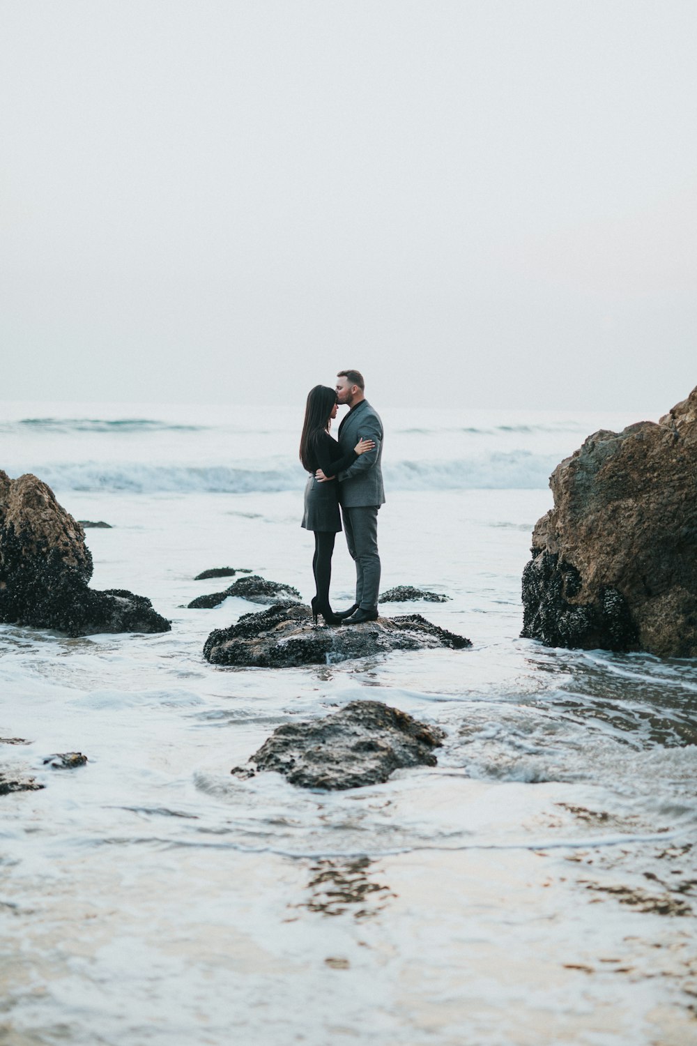 man in gray formal suit and woman in black long-sleeved top on top of rock formation beside body of water during day time