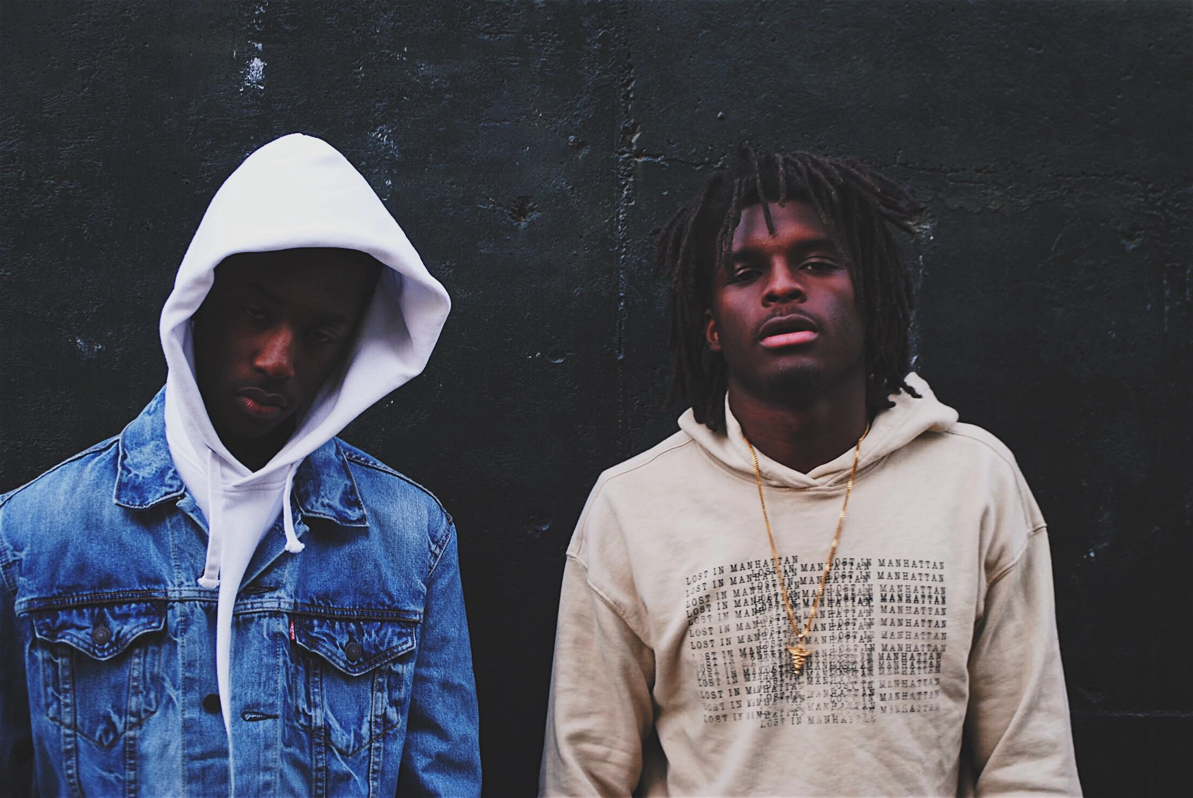 Two friends in hoodies standing against a dark wall.