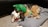 A German Shepherd dog is lying on a gray cushion with several plush toys, including a green crocodile and a white sheep. The dog appears relaxed and comfortable, surrounded by the toys.