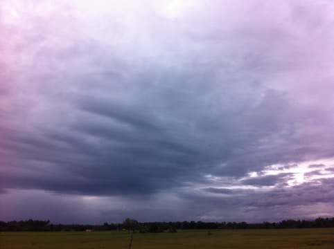 The image depicts a vast sky filled with dense, dark clouds over a flat landscape. The atmosphere appears heavy, indicative of an impending storm. The land below is a green field, possibly a rural or agricultural area, with a low, distant tree line.