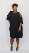 A smiling woman is standing against a plain white background, wearing a black dress with cut-out shoulders. She has curly hair and is accessorized with earrings, a bracelet, and a ring. She is posing with one hand on her hip and wearing high-heeled shoes.