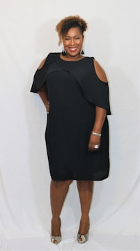 A smiling woman is standing against a plain white background, wearing a black dress with cut-out shoulders. She has curly hair and is accessorized with earrings, a bracelet, and a ring. She is posing with one hand on her hip and wearing high-heeled shoes.