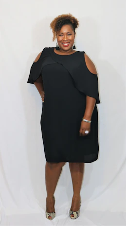 A stylish woman wearing a chic black dress with pink accessories, standing confidently against a white background.