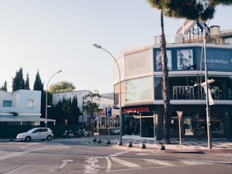 An urban street corner with a modern commercial building featuring glass facades and a visible store named 'Superdry Store'. Several storefront displays are visible, with some having posters or advertisements. There are street signs and a parking area with a few vehicles, including a white car and several scooters. Trees and green foliage are present around the buildings, with clear skies above.