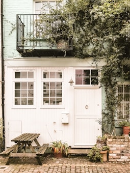 A quaint, rustic exterior of a building with a mint green brick wall. A black metal balcony is covered by leafy green vines. Below, there is a white wooden door with an accompanying window panel featuring multiple glass panes. Next to the door is a hanging light fixture and a small mailbox. In front of the building, a wooden picnic table and several potted plants are arranged on the cobblestone pavement.