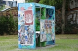 A colorful public urinal covered with various posters and graffiti stands on grass with a stone wall and greenery in the background. The structure is vibrant with blue, green, and red hues and features artistic designs alongside the posters.