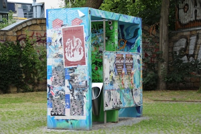 A colorful public urinal covered with various posters and graffiti stands on grass with a stone wall and greenery in the background. The structure is vibrant with blue, green, and red hues and features artistic designs alongside the posters.