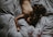 A person with long, wavy hair lies face down on a bed, surrounded by crumpled, gray sheets. The image conveys a sense of peace and relaxation.