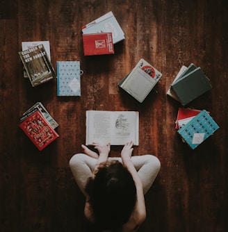 flat lay photography of woman sitting on brown wooden parquet flooring surrounded by books