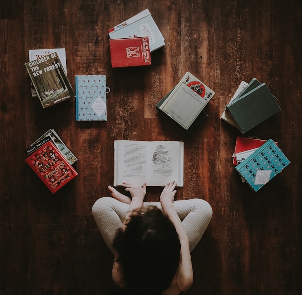 flat lay photography of woman sitting on brown wooden parquet flooring surrounded by books