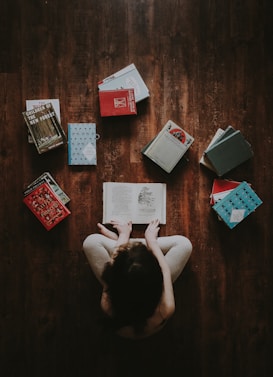 A person is sitting cross-legged on a wooden floor, surrounded by several books. The individual is reading an open book placed in their lap. The arrangement of books forms a rough circle around the reader with varied colors and sizes, creating an atmosphere of immersive reading.