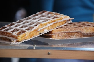 Two waffles are stacked on top of each other, dusted with powdered sugar and placed on a piece of paper. The waffles have a crisp, golden brown texture with visible grid patterns, and they rest on a reflective surface.