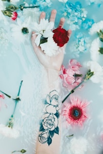 A hand with a tattoo of roses is submerged in water, surrounded by floating flowers including daisies, carnations, and chrysanthemums. The water has a milky appearance, creating a soft, tranquil atmosphere.