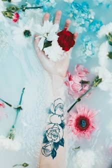 A hand with a tattoo of roses is submerged in water, surrounded by floating flowers including daisies, carnations, and chrysanthemums. The water has a milky appearance, creating a soft, tranquil atmosphere.