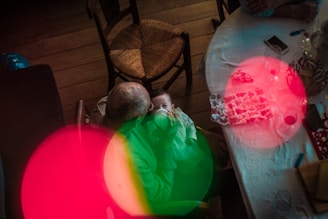 An elderly person sitting on a wooden chair holds a baby feeding from a bottle. The scene includes a table with a white tablecloth, a smartphone, cups, and wrapped gifts. Bright red and green light reflections add a dreamy effect.