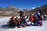 A group of people is gathered on a snowy surface in a mountainous region. They are all dressed in winter clothing, with some wearing hats and sunglasses. The background features majestic mountain peaks under a clear blue sky.